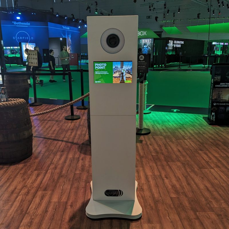 A free-standing white photobooth tower with a ring-lit camera and touchscreen on an exhibition stand