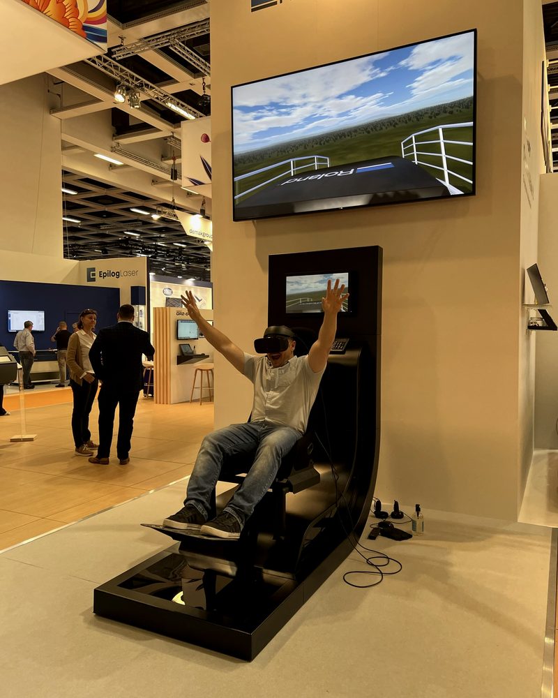 A rider in a VR headset with arms raised on the full-motion roller coaster, the ride shown on a large screen above